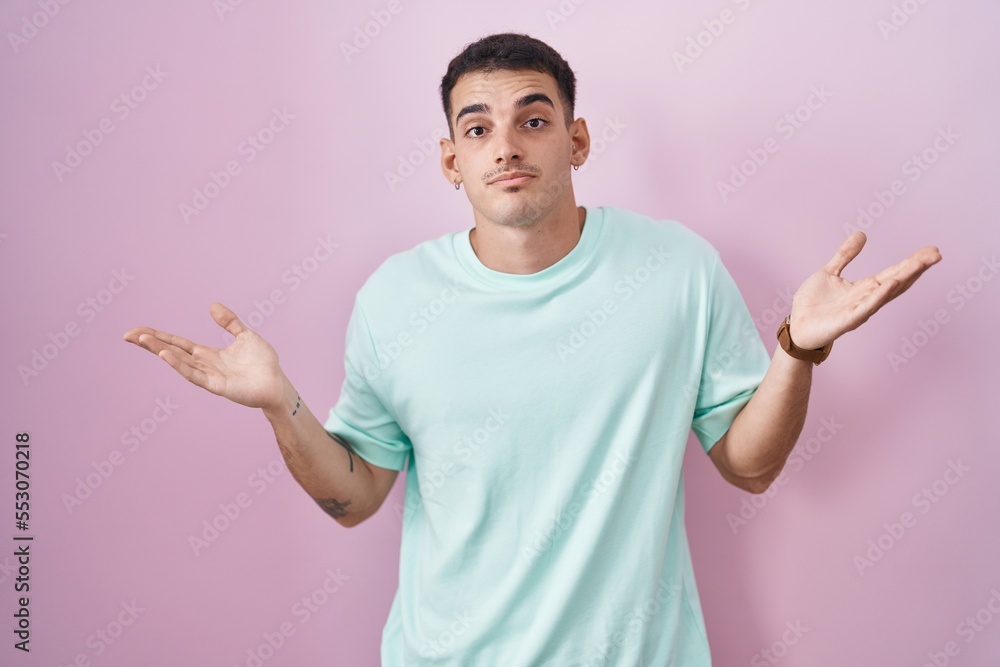 Handsome hispanic man standing over pink background clueless and confused expression with arms and hands raised. doubt concept. Handsome hispanic man standing over pink background clueless and confused expression with arms and hands raised. doubt concept.