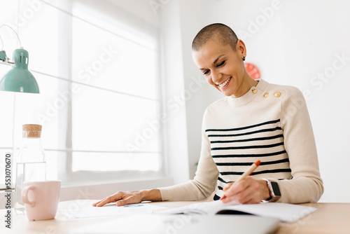 Positive mid adult businesswoman working from home office. Successful female entrepreneur writing on notebook sitting at workplace