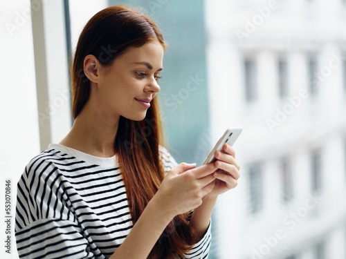 Woman holding her phone and looking and smiling at it near the window of the house, online shopping from home