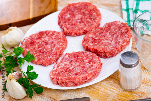 Shaped patties for burgers from raw ground beef meat with fresh parsley sprigs, spicy garlic and allspice on plate. Cooking ingredients
