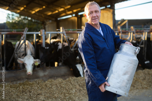 Elderly male owner with milk can standing in stall on background with herd of cows on farm