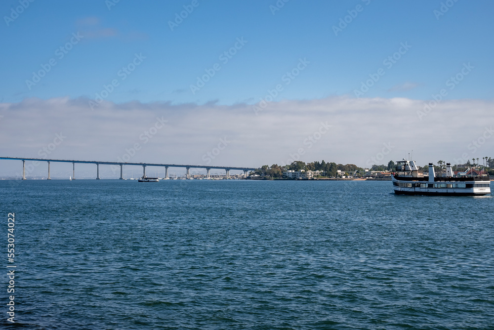 Ship sailing on San Diego bay and distant view of Coronado bridge over seascape with cloudy sky in the background during sunny day