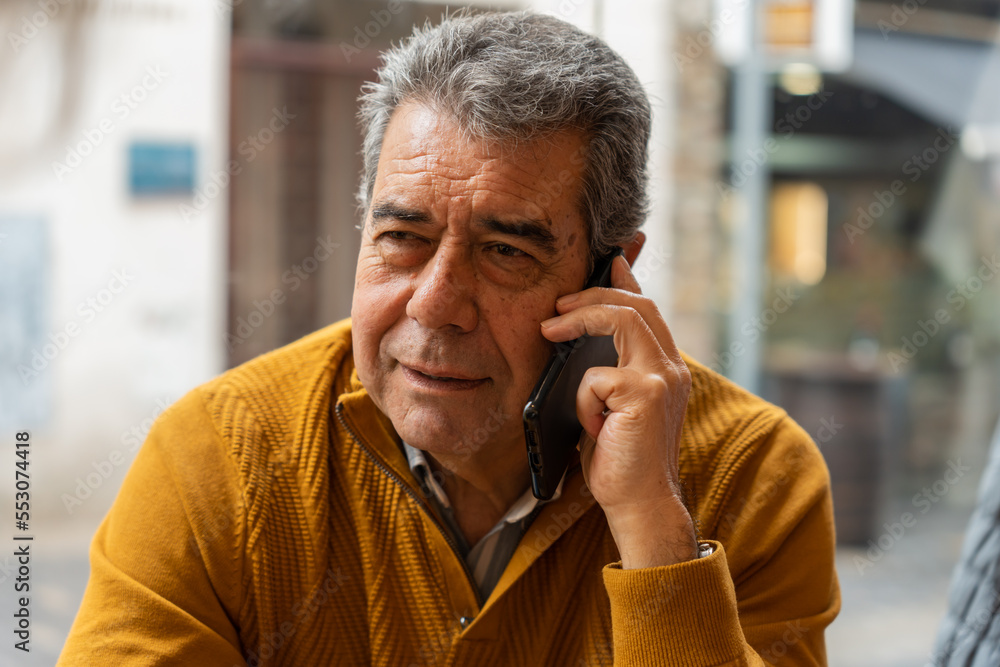 Middle aged man with gray hair looking away and answering phone call on blurred background of city street Middle aged man with gray hair looking away and answering phone call on blurred background of city street