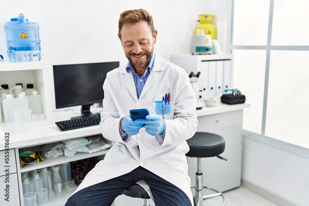 Middle age hispanic man wearing scientist uniform using smartphone at laboratory Middle age hispanic man wearing scientist uniform using smartphone at laboratory