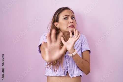 Young hispanic woman standing over pink background afraid and terrified with fear expression stop gesture with hands  shouting in shock. panic concept.