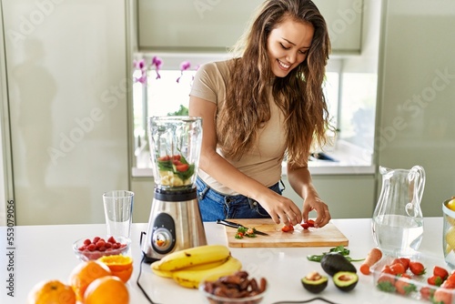 Young beautiful hispanic woman preparing vegetable smoothie with blender at the kitchen