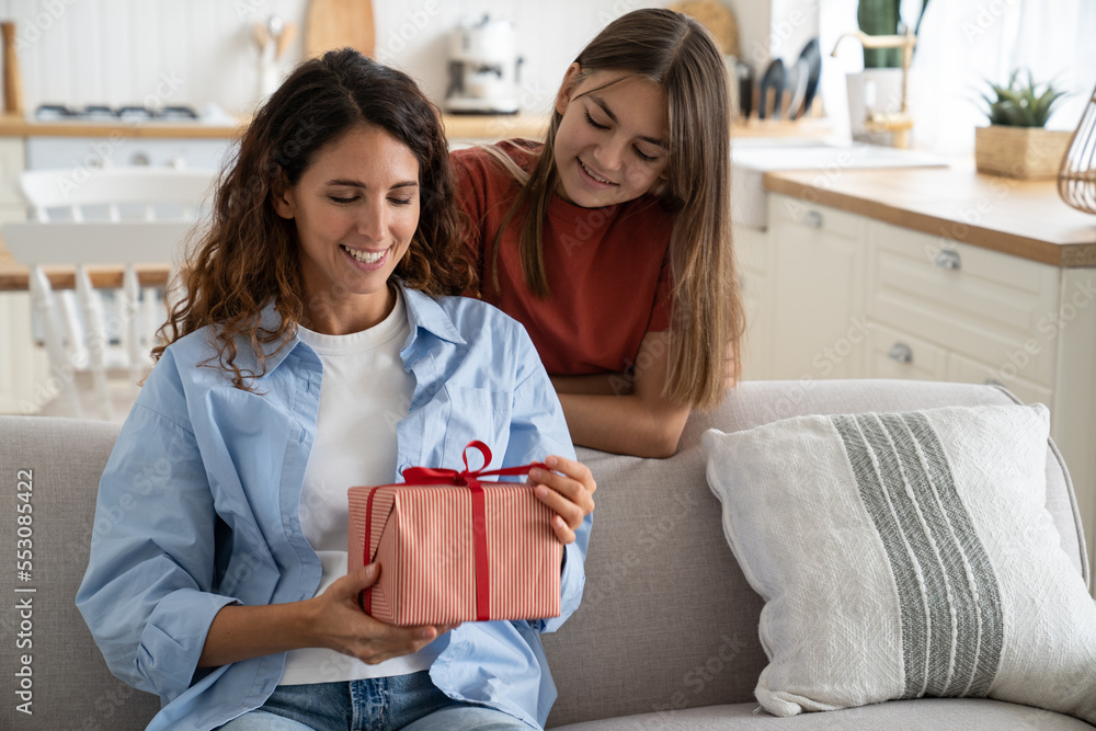 Young happy curious woman mother sitting on couch at home holding wrapped gift box from daughter, opening present. Smiling little girl kid making surprise to mom. Mothers day celebration concept Young happy curious woman mother sitting on couch at home holding wrapped gift box from daughter, opening present. Smiling little girl kid making surprise to mom. Mothers day celebration concept