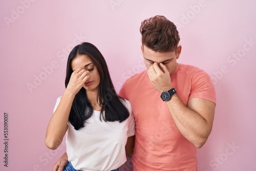 Young hispanic couple standing over pink background tired rubbing nose and eyes feeling fatigue and headache. stress and frustration concept.