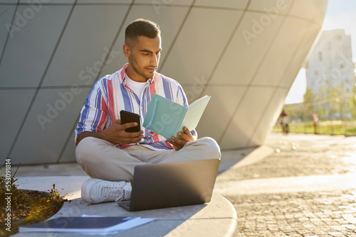 Pensive middle eastern student studying, exam preparation, reading book in university campus. Modern Iranian man using laptop, holding mobile phone working sitting at workplace 