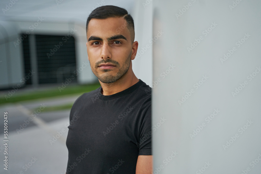 Portrait of young serious Iranian man with stylish hair cut wearing black t shirt looking at camera on the street, copy space Portrait of young serious Iranian man with stylish hair cut wearing black t shirt looking at camera on the street, copy space