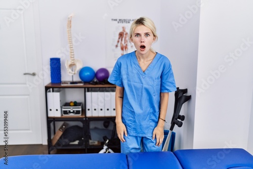 Young physiotherapist woman working at pain recovery clinic afraid and shocked with surprise and amazed expression, fear and excited face.