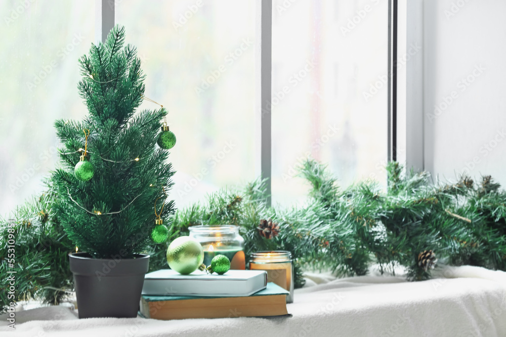Small Christmas tree with balls, books, candles and fir branches on windowsill in room