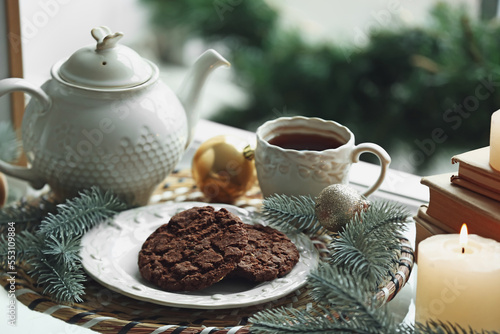 Plate with cookies, teapot, cup of tea, Christmas branches and balls on windowsill, closeup