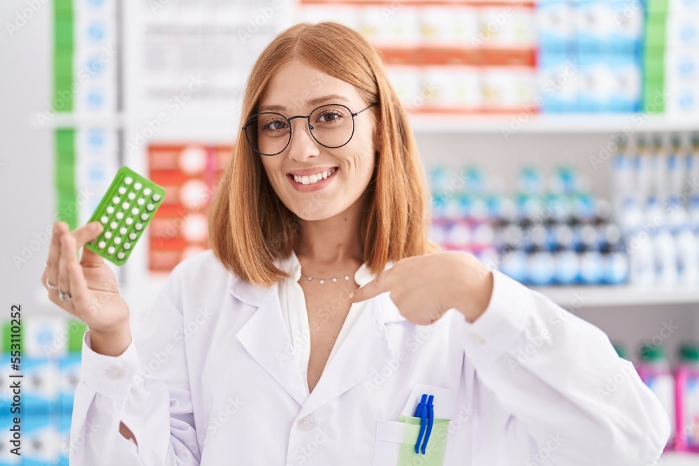 Young redhead woman working at pharmacy drugstore holding birth control pills pointing finger to one self smiling happy and proud