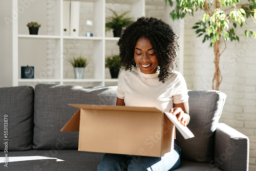 Young african woman sit on couch at home unpacking parcel cardboard box with online purchase