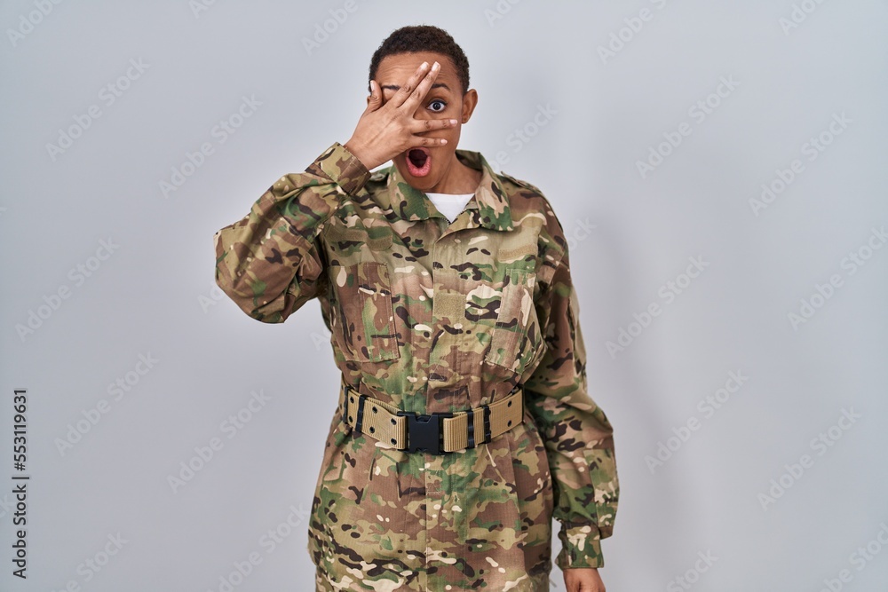 Beautiful african american woman wearing camouflage army uniform peeking in shock covering face and eyes with hand, looking through fingers with embarrassed expression. Beautiful african american woman wearing camouflage army uniform peeking in shock covering face and eyes with hand, looking through fingers with embarrassed expression.