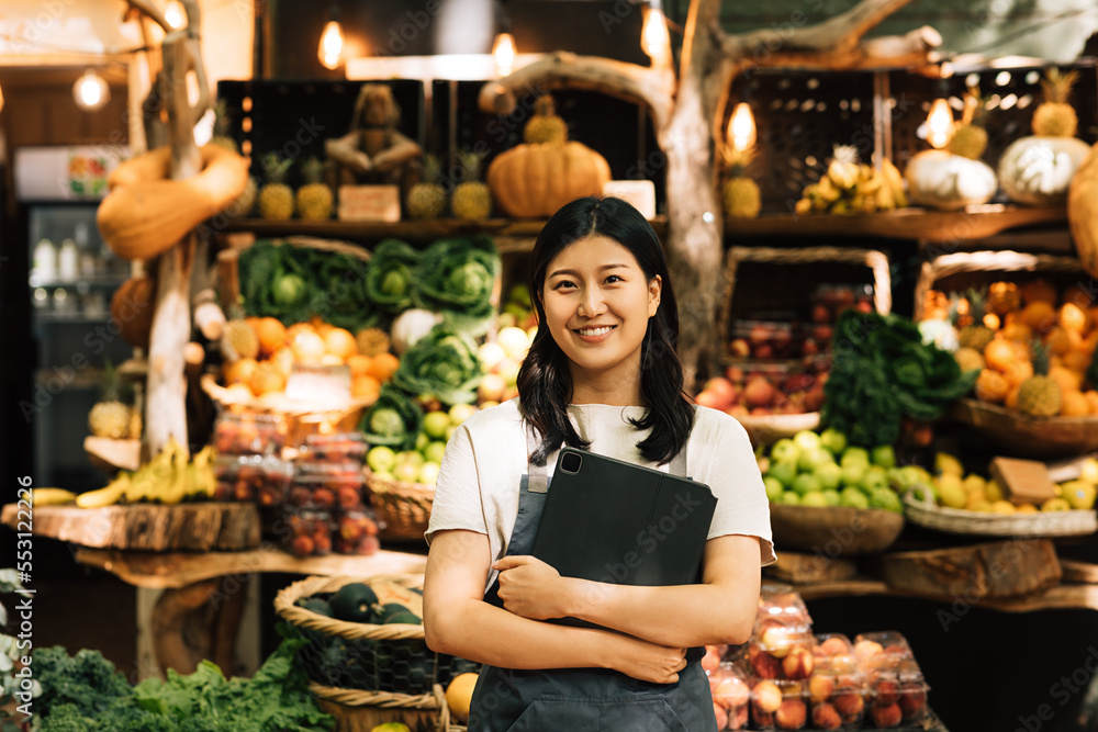 Smiling entrepreneur holding a digital tablet standing at an outdoor market. Asian woman in an apron looking at camera while standing against a street food market. - obrazy, fototapety, plakaty 