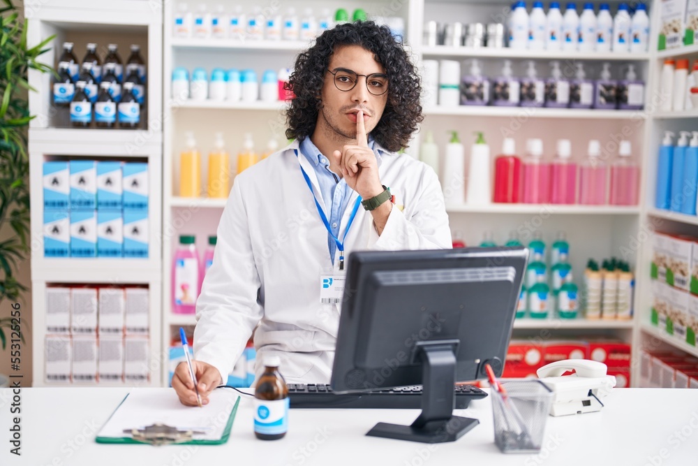 Hispanic man with curly hair working at pharmacy drugstore asking to be quiet with finger on lips. silence and secret concept.