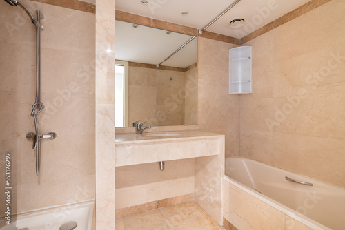 Bright bathroom with tiled walls and floors in warm light beige tones. Washbasin with large mirror  white acrylic bathtub and shower area for taking morning shower everything you need is here.