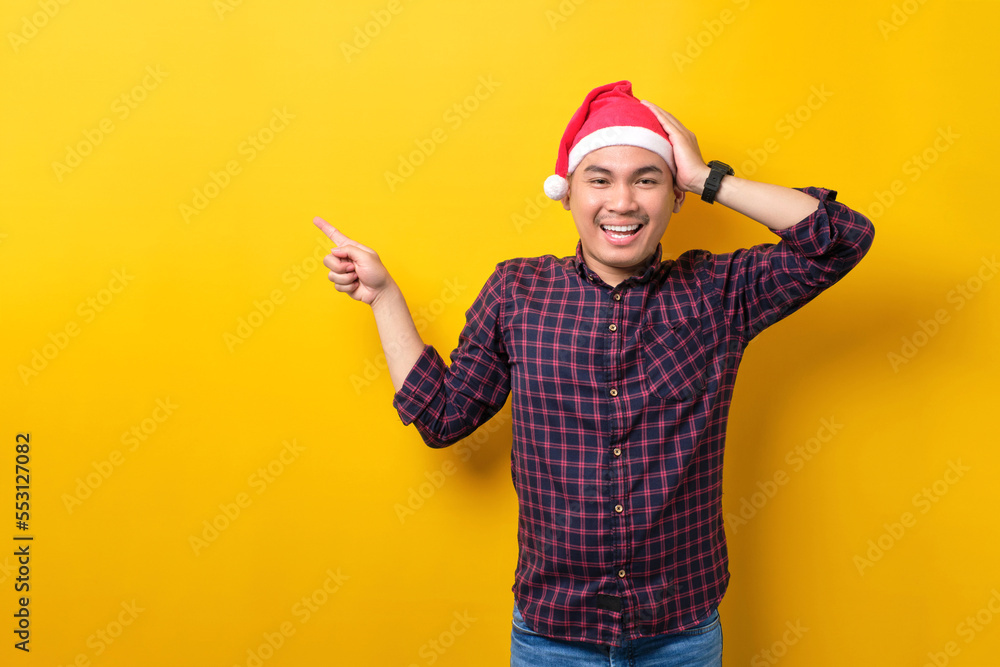 Cheerful young Asian man in Santa hat pointing fingers aside at blank space for advertising text on yellow studio background. Happy New Year 2023 celebration merry holiday concept