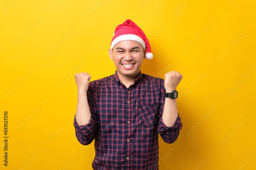Excited young Asian man in Santa hat raising hands up, celebrating success on yellow studio background. Happy New Year 2023 celebration merry holiday concept Excited young Asian man in Santa hat raising hands up, celebrating success on yellow studio background. Happy New Year 2023 celebration merry holiday concept