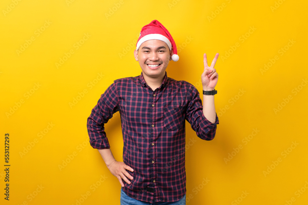 Cheerful young Asian man in Santa hat showing victory sign, looking at camera on yellow studio background. Happy New Year 2023 celebration merry holiday concept
