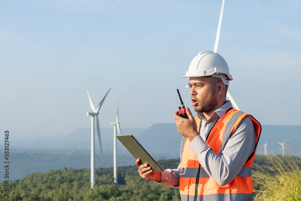 Portrait of handsome asian man engineer wearing safety helmet and reflective vest talking to his team or someone with radio communication with wind turbine propeller background. Portrait of handsome asian man engineer wearing safety helmet and reflective vest talking to his team or someone with radio communication with wind turbine propeller background.