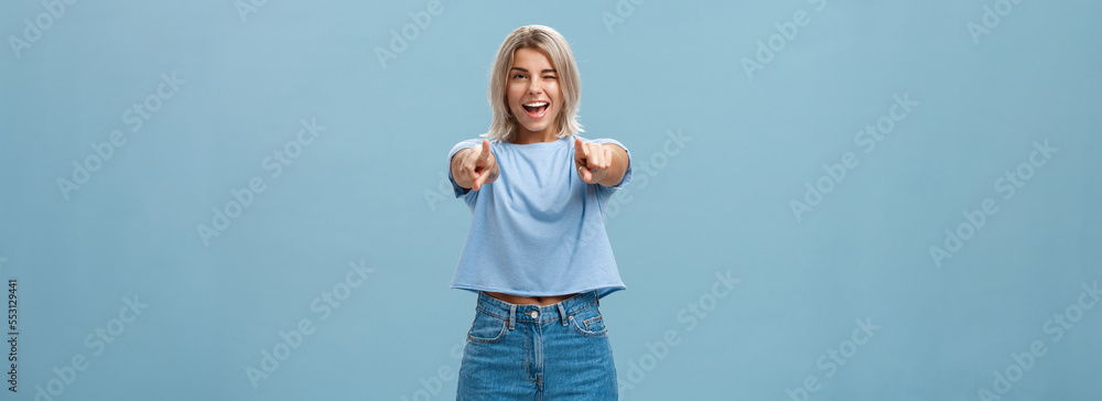 Happy enthusiastic stylish blonde female with tattoos pointing at camera as if picking or making hint smiling joyfully and winking from amazement and happiness posing over blue background - obrazy, fototapety, plakaty 
