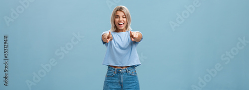 Happy enthusiastic stylish blonde female with tattoos pointing at camera as if picking or making hint smiling joyfully and winking from amazement and happiness posing over blue background