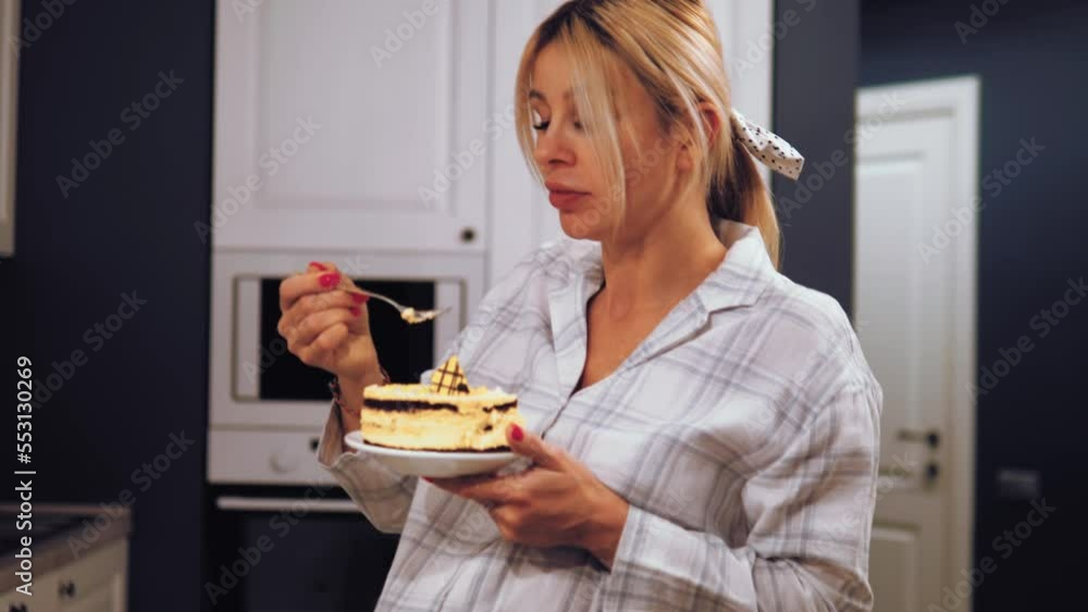 Happy pregnant woman eating a sweet cake. Pregnant girl eating cake at home. Harmful food during pregnancy. Pregnant Female enjoying sweet dessert cake standing in the kitchen Happy pregnant woman eating a sweet cake. Pregnant girl eating cake at home. Harmful food during pregnancy. Pregnant Female enjoying sweet dessert cake standing in the kitchen