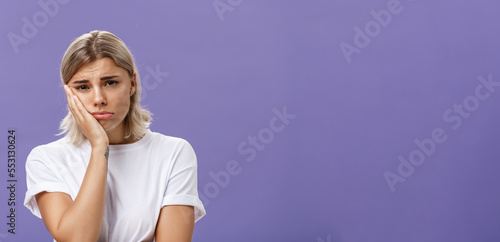 Waist-up shot of unhappy miserable and sad cute blonde female in white casual t-shirt pursing lips leaning face on palm and frowning from disappointement and regret over purple background photo