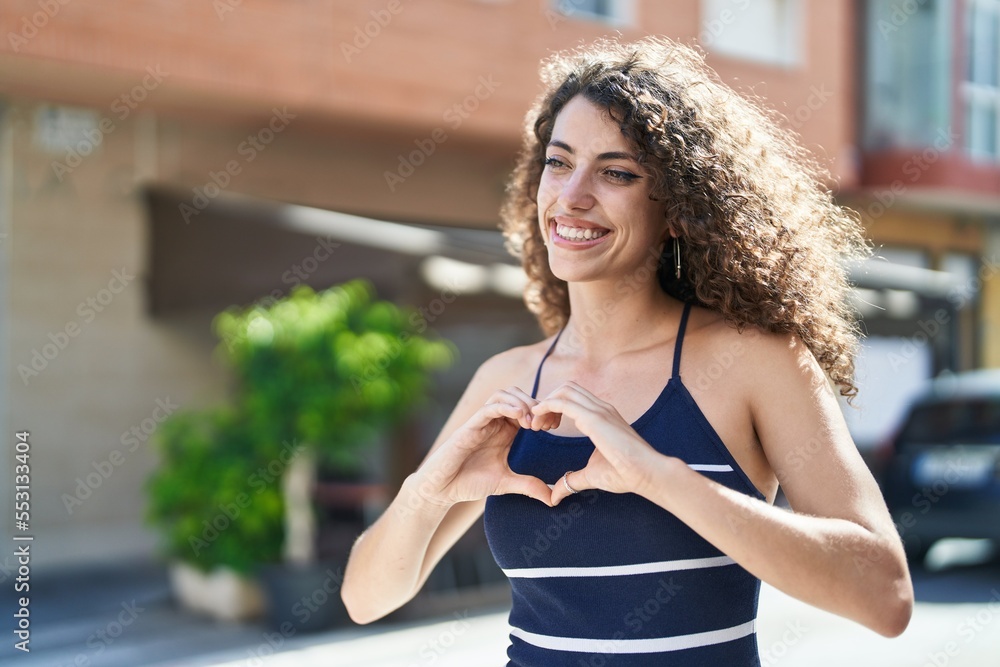 Young beautiful hispanic woman smiling confident doing heart gesture with hands at street