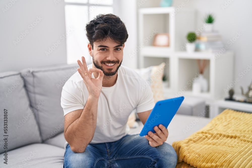 Hispanic man with beard using touchpad sitting on the sofa smiling positive doing ok sign with hand and fingers. successful expression.