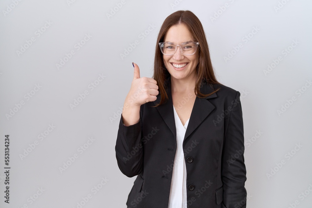 Beautiful brunette woman wearing business jacket and glasses doing happy thumbs up gesture with hand. approving expression looking at the camera showing success. Beautiful brunette woman wearing business jacket and glasses doing happy thumbs up gesture with hand. approving expression looking at the camera showing success.