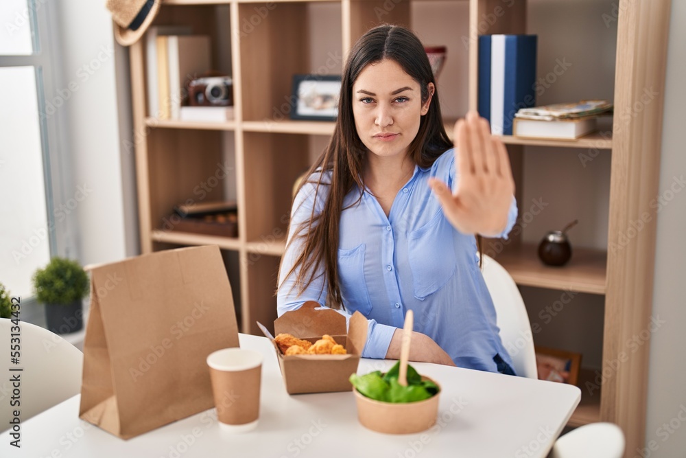 Young brunette woman eating take away food at home with open hand doing stop sign with serious and confident expression, defense gesture Young brunette woman eating take away food at home with open hand doing stop sign with serious and confident expression, defense gesture