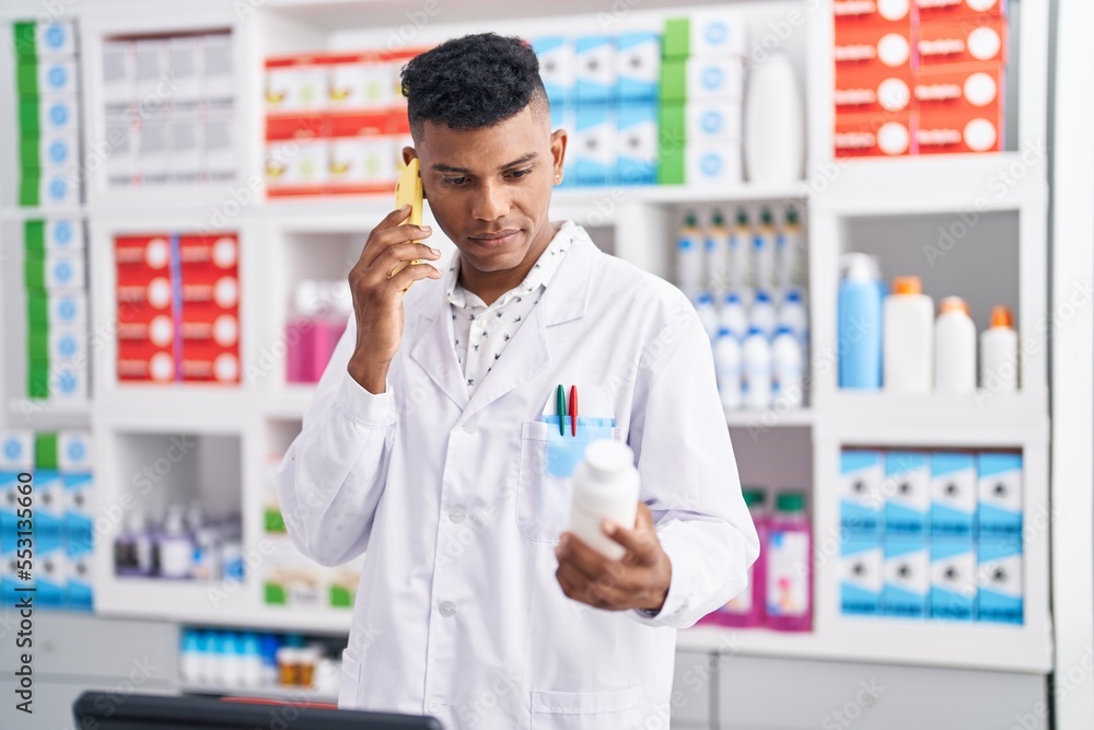 Young latin man pharmacist talking on smartphone holding pills bottle at pharmacy