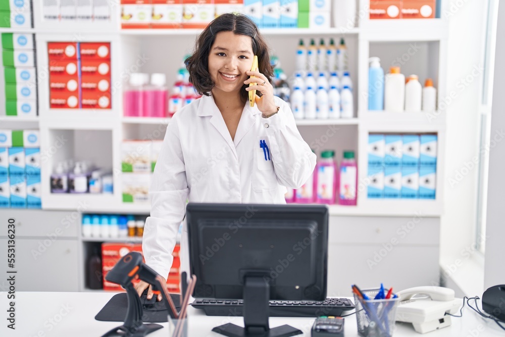 Young woman pharmacist talking on smartphone using computer at pharmacy Young woman pharmacist talking on smartphone using computer at pharmacy