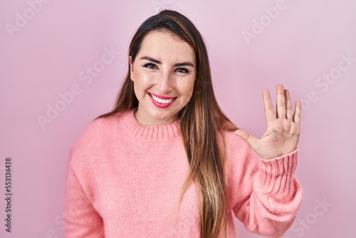 Young hispanic woman standing over pink background showing and pointing up with fingers number five while smiling confident and happy.