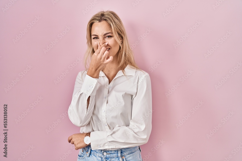 Young caucasian woman wearing casual white shirt over pink background smelling something stinky and disgusting, intolerable smell, holding breath with fingers on nose. bad smell
