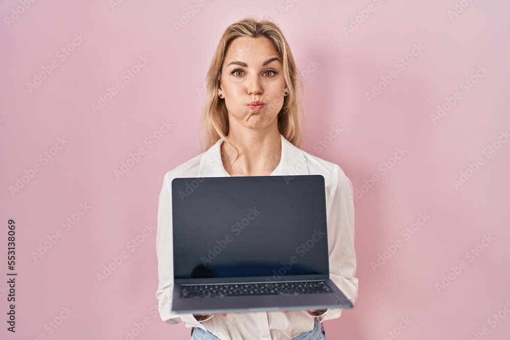 Young caucasian woman holding laptop showing screen puffing cheeks with funny face. mouth inflated with air, catching air. - obrazy, fototapety, plakaty 