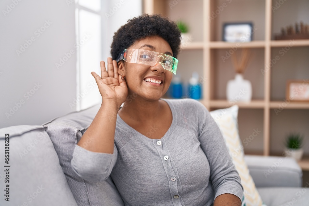 Young african american woman wearing virtual reality glasses sitting on the sofa smiling with hand over ear listening and hearing to rumor or gossip. deafness concept. Young african american woman wearing virtual reality glasses sitting on the sofa smiling with hand over ear listening and hearing to rumor or gossip. deafness concept.