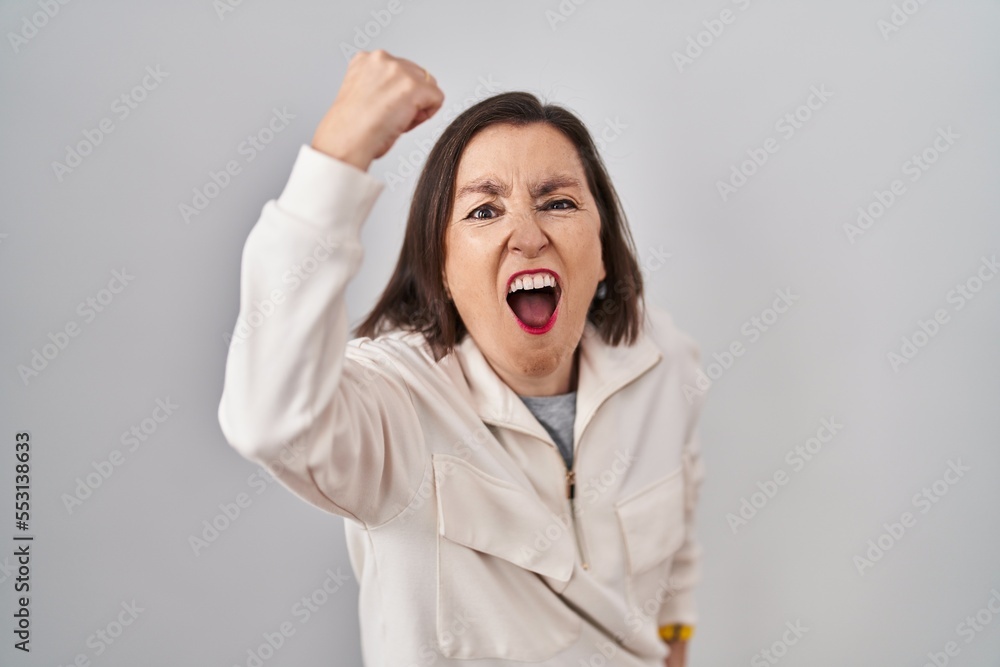 Middle age hispanic woman standing over isolated background angry and mad raising fist frustrated and furious while shouting with anger. rage and aggressive concept. Middle age hispanic woman standing over isolated background angry and mad raising fist frustrated and furious while shouting with anger. rage and aggressive concept.