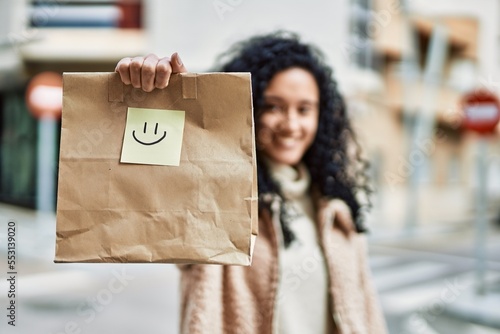Young hispanic woman smiling confident holding take away paper bag at street