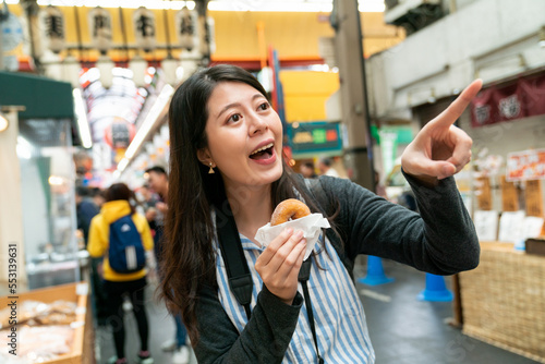 excited asian female traveler noticing something fun and finger pointing at distance while having baked doughnut for snack in kuromon ichiba market in Osaka japan