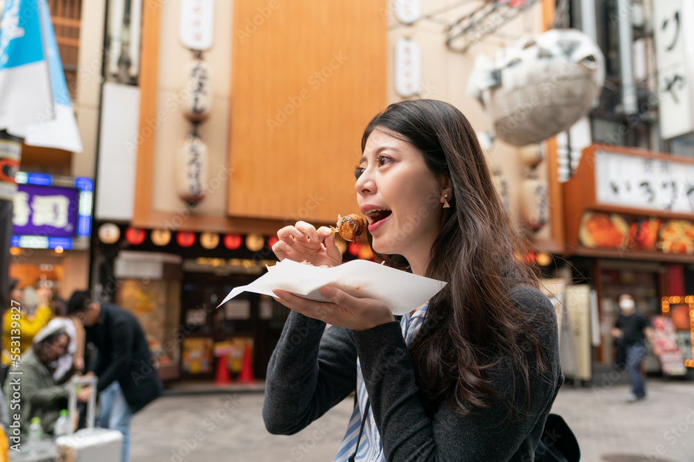 side view of cheerful Asian woman visitor eating takoyaki octopus ball while taking leisure walk on the shopping district in shinsaibashisuji and doutonbori Osaka japan