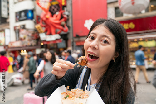 pretty happy Asian woman visitor looking at camera and enjoying tasty street food takoyaki with a pick in shinsaibashisuji and doutonbori shopping area Osaka japan