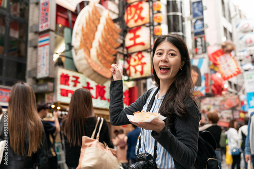 amazed Asian female visitor holding takoyaki and pointing at giant dumpling sign at background while visiting shinsaibashisuji and doutonbori Osaka japan. translation: âdumpling shopâ
