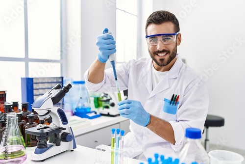 Young hispanic man wearing scientist uniform using pipette and test tube at laboratory
