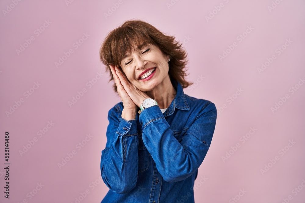Middle age woman standing over pink background sleeping tired dreaming and posing with hands together while smiling with closed eyes. Middle age woman standing over pink background sleeping tired dreaming and posing with hands together while smiling with closed eyes.