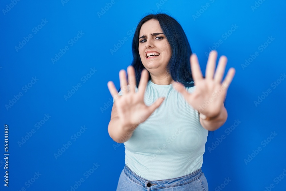 Fototapeta premium Young modern girl with blue hair standing over blue background afraid and terrified with fear expression stop gesture with hands, shouting in shock. panic concept.